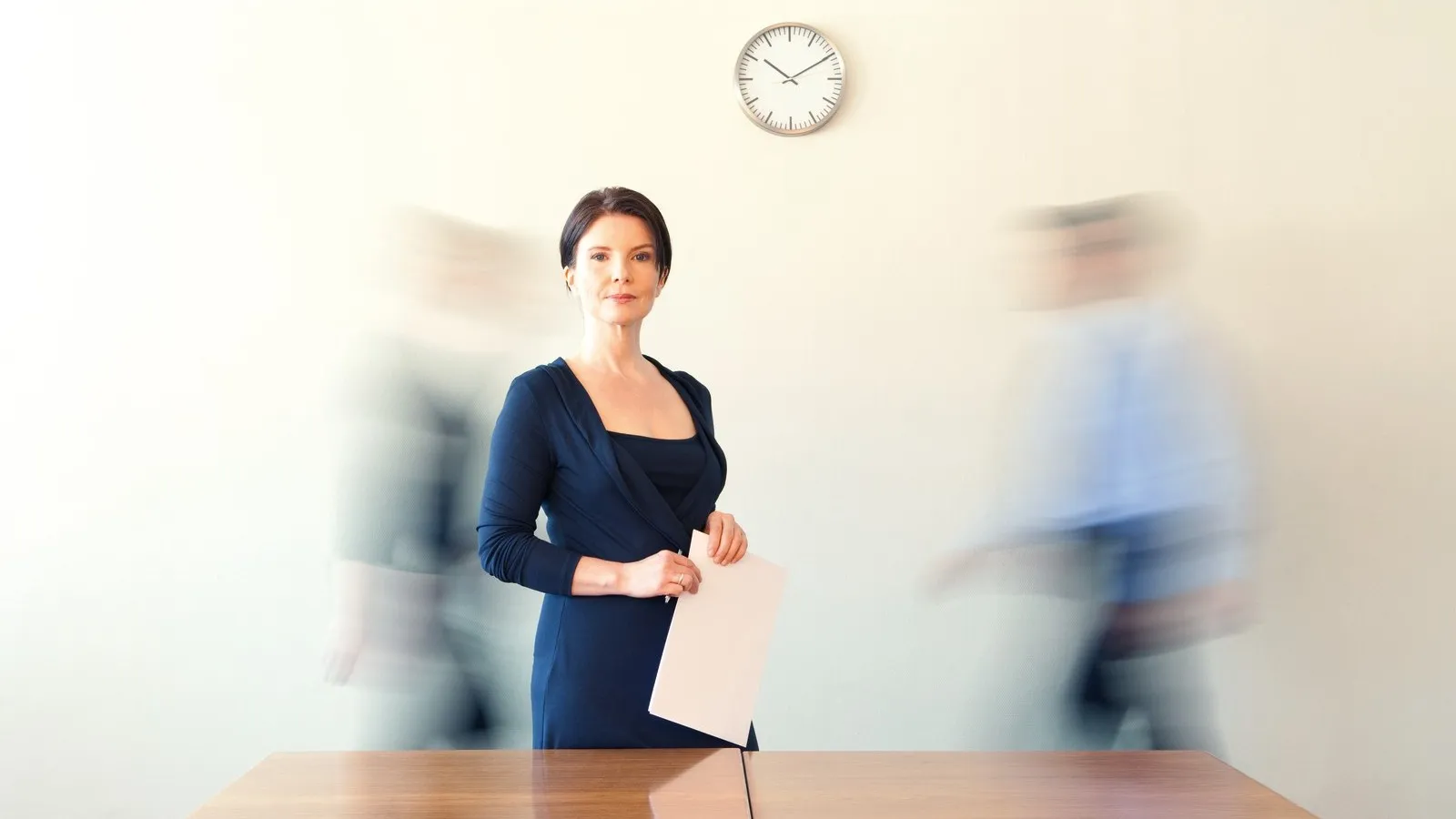 Eine Frau steht im Buero am Konferenztisch, zwei Kollegen in Bewegung. An der Wand hängt mittig eine Uhr.